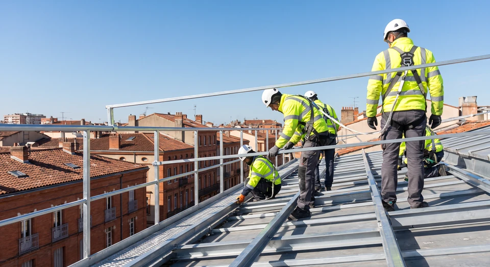 Photographie réaliste d'un chantier de construction ou de rénovation en hauteur : plusieurs ouvriers équipés de gilets de sécurité travaillent sur une structure métallique ou un échafaudage. Au premier plan, un garde-corps industriel ou une barrière de sécurité métallique bien visible protège la zone de travail en hauteur. La photo doit montrer clairement le matériel de sécurité en place, l'environnement de chantier professionnel, et l'équipe au travail pour incarner l'enjeu de sécurité pour les équipes sur le terrain.