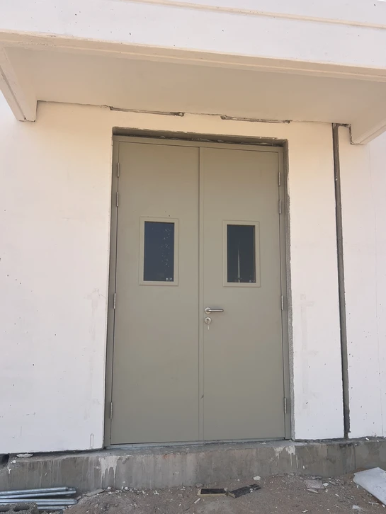 Gros plan d'un technicien en tenue de travail professionnelle (combinaison ou salopette) installant ou travaillant sur une porte coupe-feu métallique robuste dans un bâtiment commercial ou industriel. La porte présente les caractéristiques typiques d'une porte coupe-feu : cadre métallique renforcé, joints d'étanchéité visibles, quincaillerie de sécurité. L'environnement montre un contexte professionnel B2B (bâtiment commercial, industriel ou immeuble). L'action illustre la rigueur technique et la précision de l'installation, en accord avec le parcours aéronautique du dirigeant.